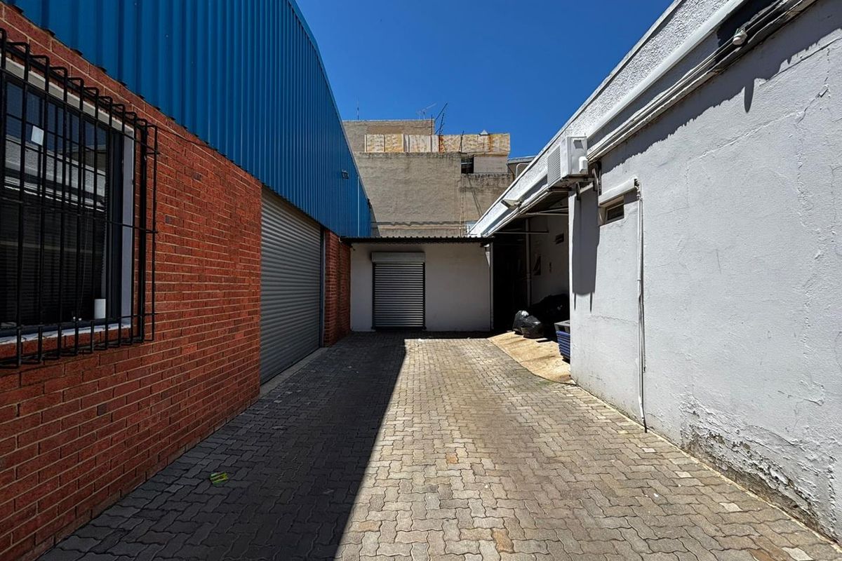 A paved rear service alleyway between two industrial buildings, featuring roller shutter access doors and security-barred windows.