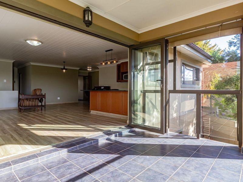 A covered balcony with tiled flooring and glass balustrades, featuring seamless access to an open-plan living and kitchen area with laminate flooring.