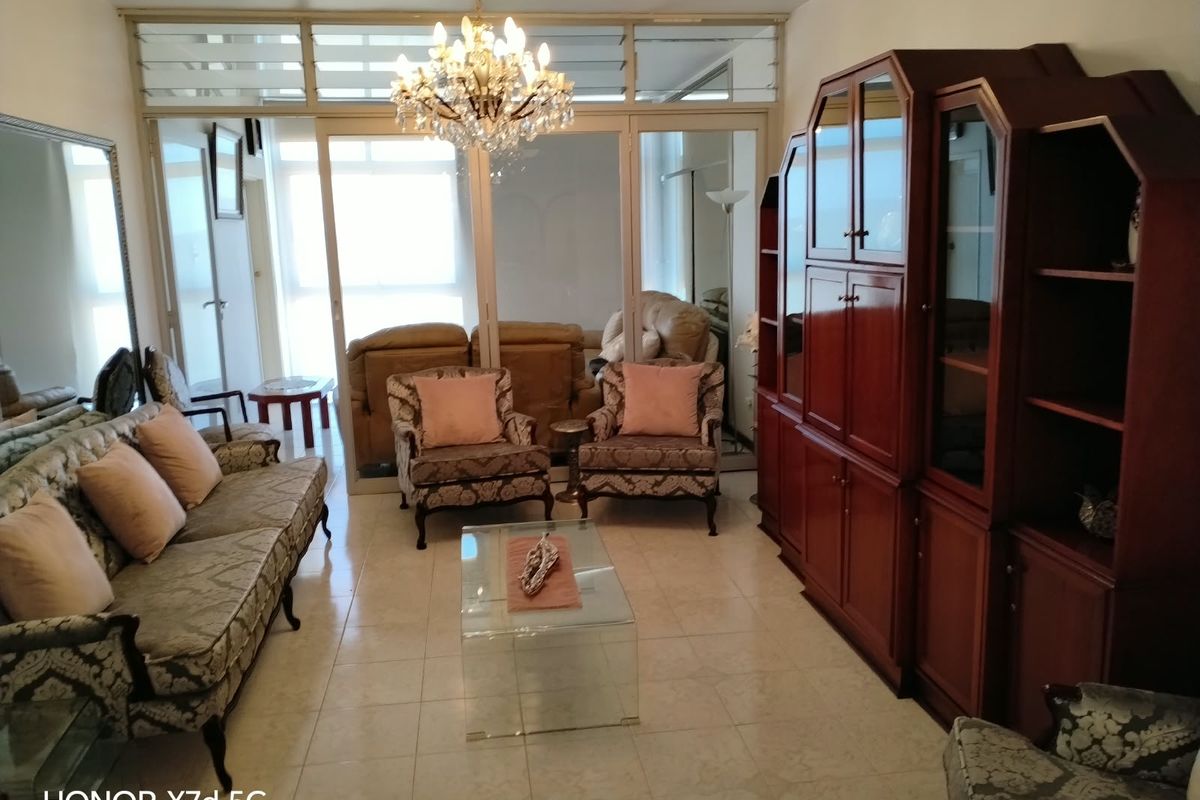 A spacious living room featuring tiled flooring, a crystal chandelier, and large glass sliding doors that provide natural light and access to the exterior.