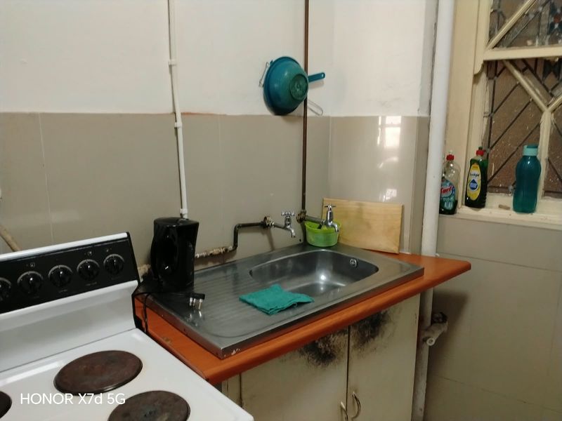 A compact kitchen area featuring a stainless steel sink, tiled splashback, and built-in cabinetry with visible signs of wear and exposed plumbing.