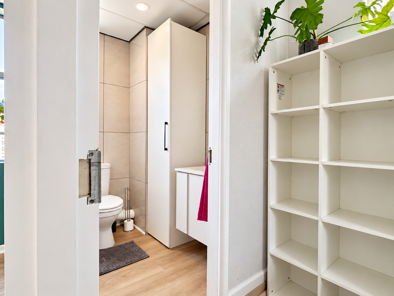 A clean and functional bathroom featuring a built-in vanity, tall storage cabinetry, and neutral wall tiling, accessible from an adjacent hallway area.