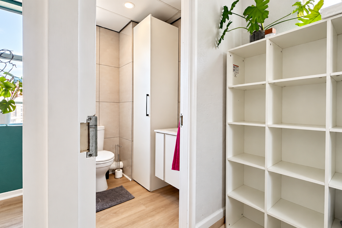 A clean and functional bathroom featuring a built-in vanity, tall storage cabinetry, and neutral wall tiling, accessible from an adjacent hallway area.