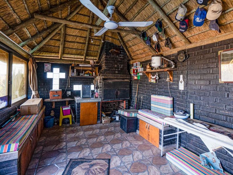An enclosed patio area featuring a traditional thatched roof, exposed timber rafters, built-in brick braai, and integrated bench seating with tiled flooring.