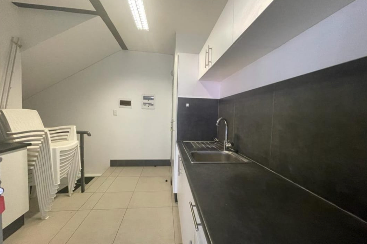 A compact kitchenette area featuring a stainless steel sink, dark countertops, and white cabinetry, set within an office space with tiled flooring.