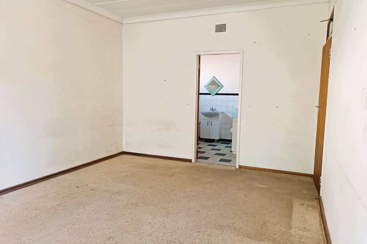 An empty bedroom featuring worn carpeting and plain walls, with an adjoining bathroom visible through the doorway.