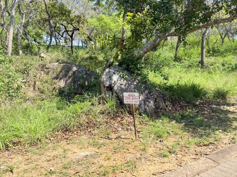 A vacant residential plot featuring natural rock formations and mature trees, situated adjacent to a paved street verge.