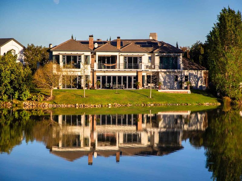 A stunning rear view of a two-story luxury residence featuring a tiled roof, solar panels, and expansive glass doors overlooking a tranquil waterfront.