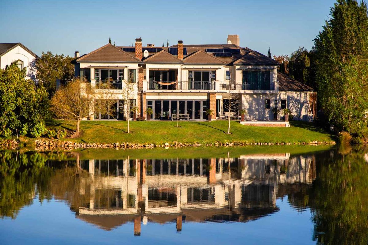 A stunning rear view of a two-story luxury residence featuring a tiled roof, solar panels, and expansive glass doors overlooking a tranquil waterfront.