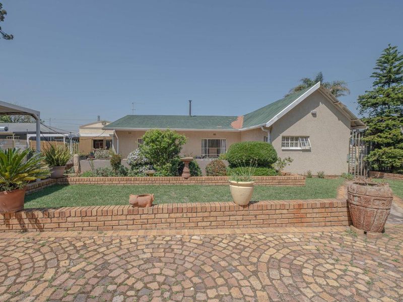 Rear view of the property featuring a landscaped garden, brick retaining walls, and cobblestone paving with a metal roof structure.