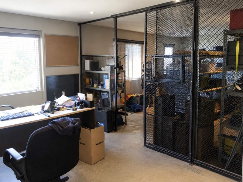 Industrial office space featuring a secure wire mesh partition, concrete flooring, and natural light through venetian blinds, suitable for a workshop or storage-integrated workspace.