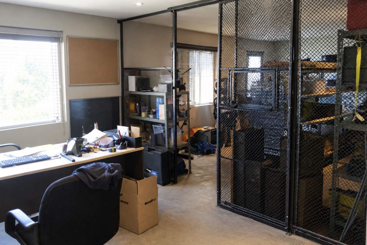 Industrial office space featuring a secure wire mesh partition, concrete flooring, and natural light through venetian blinds, suitable for a workshop or storage-integrated workspace.
