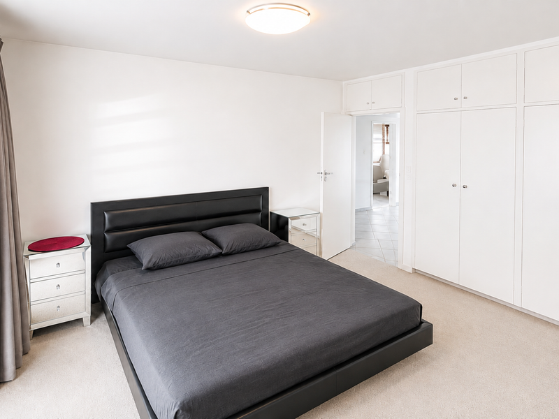 A well-maintained bedroom featuring neutral carpeting, ample built-in cupboard storage, and a bright, clean aesthetic with direct access to an adjoining hallway.