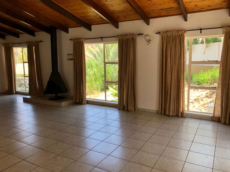 Spacious living area featuring exposed timber ceiling beams, tiled flooring, and a freestanding fireplace with large windows overlooking the garden.