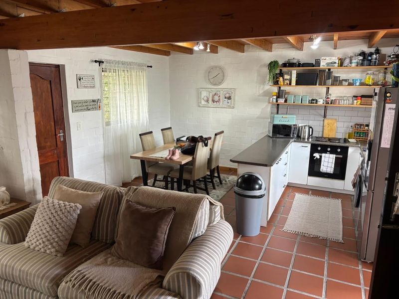 Open-plan living and kitchen area featuring exposed timber beams, painted brick walls, terracotta flooring, and a functional kitchen with built-in cabinetry and shelving.