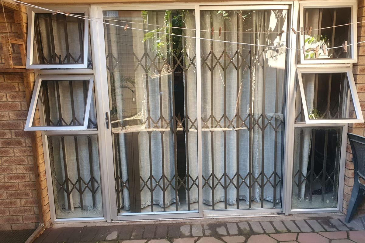 Rear exterior view featuring a sliding glass door with security gate, flanked by top-hung windows and set against a brick facade with paved patio.