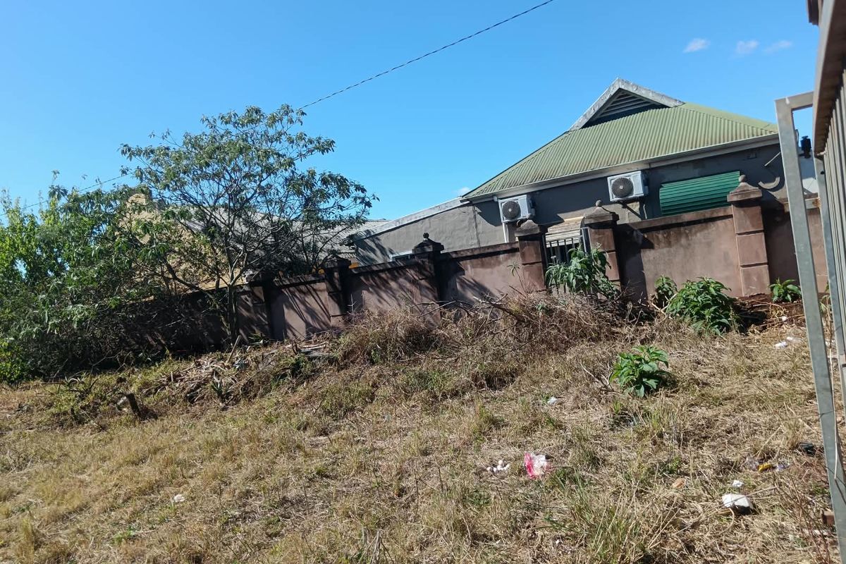 A view of the rear boundary wall and adjacent property structure, featuring masonry pillars and mounted air conditioning units on a sloped, overgrown plot.