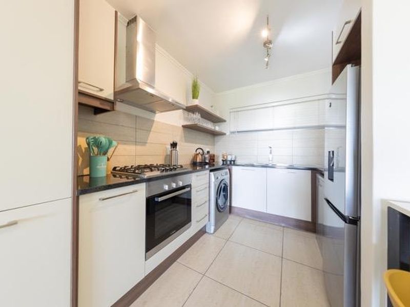 Modern galley-style kitchen featuring sleek cabinetry, built-in oven, gas hob, stainless steel extractor fan, and integrated laundry appliances with tiled flooring.