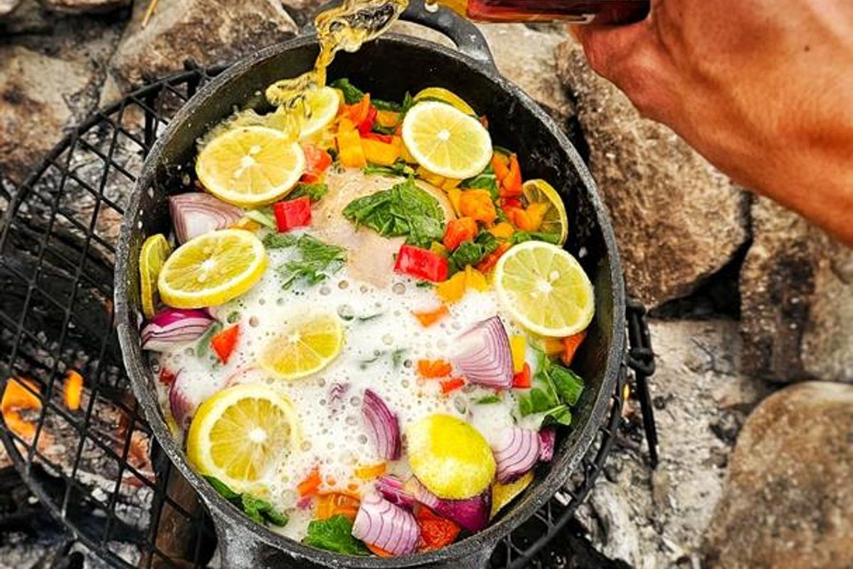 An outdoor cooking scene featuring a cast iron pot over a fire pit, being prepared with fresh vegetables and citrus.
