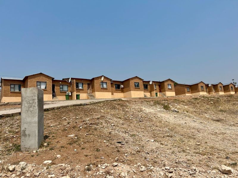 A row of single-story brick townhouse units featuring concrete driveways and exterior-mounted air conditioning units, set against an unlandscaped, rocky terrain.