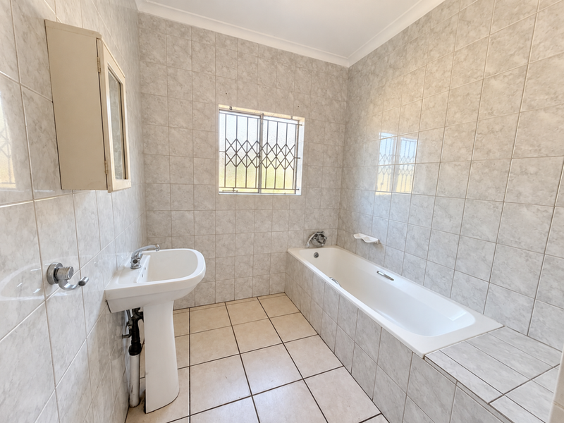 A functional bathroom featuring a pedestal basin, a built-in bathtub, and full-height wall tiling with a secure window.