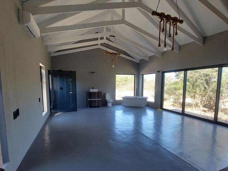 Spacious open-plan bathroom featuring vaulted ceilings with exposed rafters, polished concrete floors, a freestanding bathtub, walk-in shower, and large sliding doors with natural views.