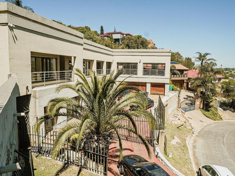 Elevated view of the property front facade featuring a double garage, paved driveway, security fencing, and an upper-level balcony with sliding glass doors.