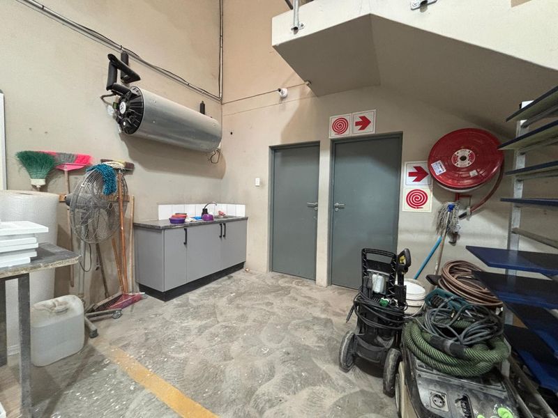 Industrial utility space featuring a kitchenette cabinet, sink, wall-mounted water heater, and fire safety equipment, with concrete flooring and access doors.