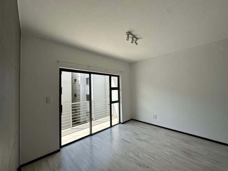 A bright, empty living room featuring modern laminate flooring, a ceiling-mounted spotlight track, and large sliding glass doors leading to a private balcony.