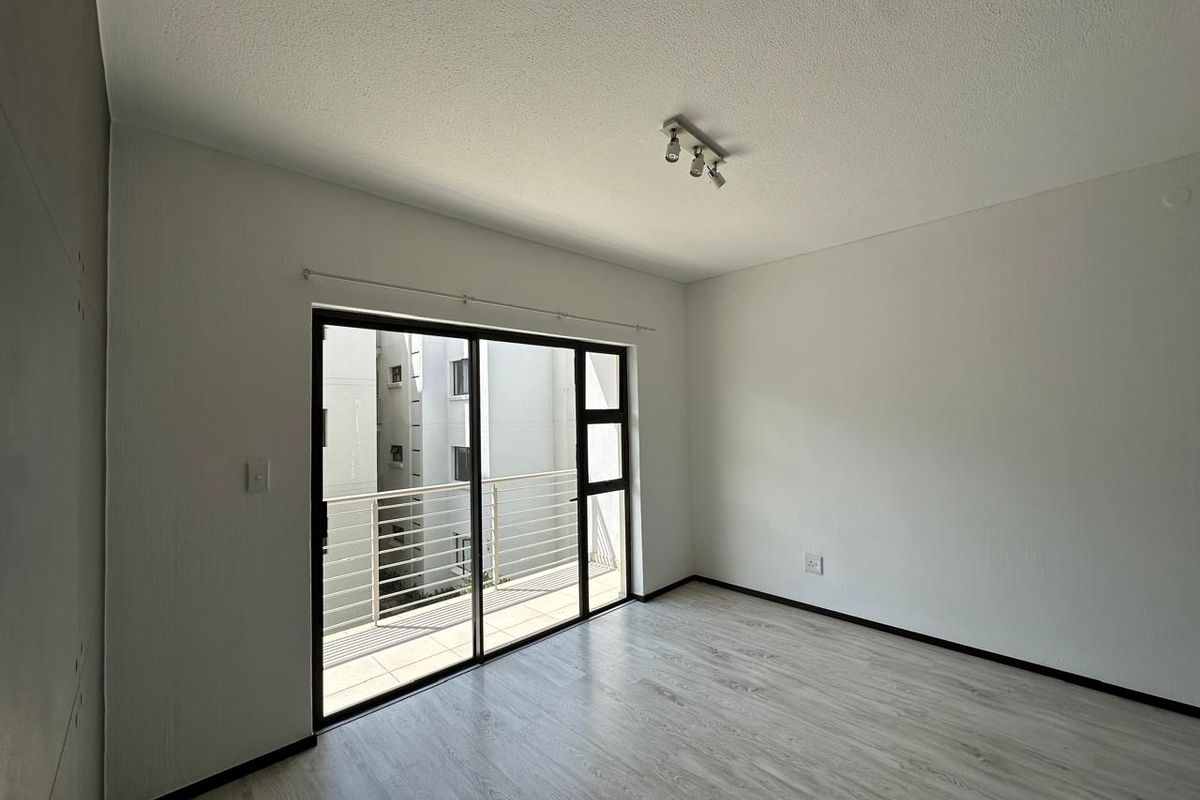 A bright, empty living room featuring modern laminate flooring, a ceiling-mounted spotlight track, and large sliding glass doors leading to a private balcony.