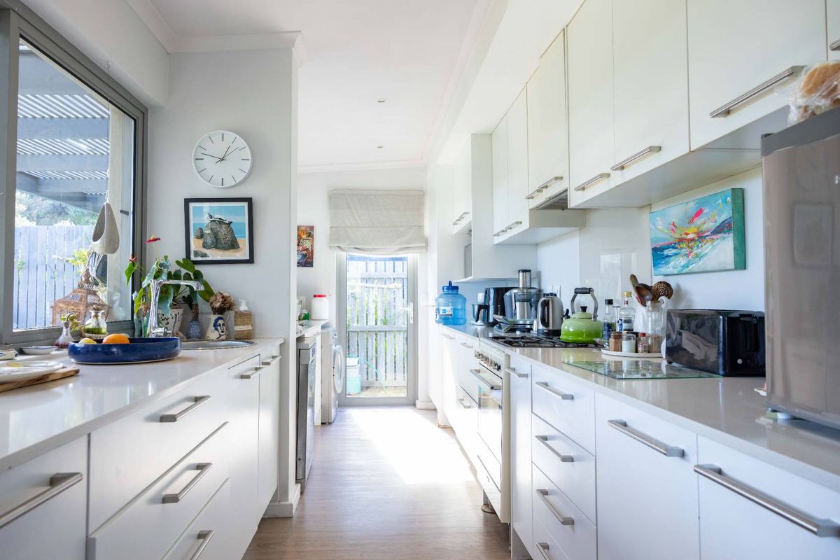 A bright, modern galley-style kitchen featuring white cabinetry, stone countertops, integrated appliances, and direct access to the rear exterior.