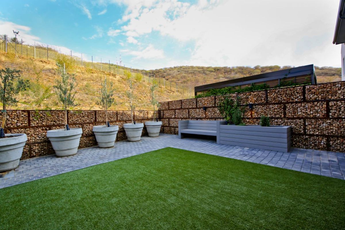 A well-maintained rear garden featuring a paved patio area, artificial lawn, and decorative gabion stone retaining walls.