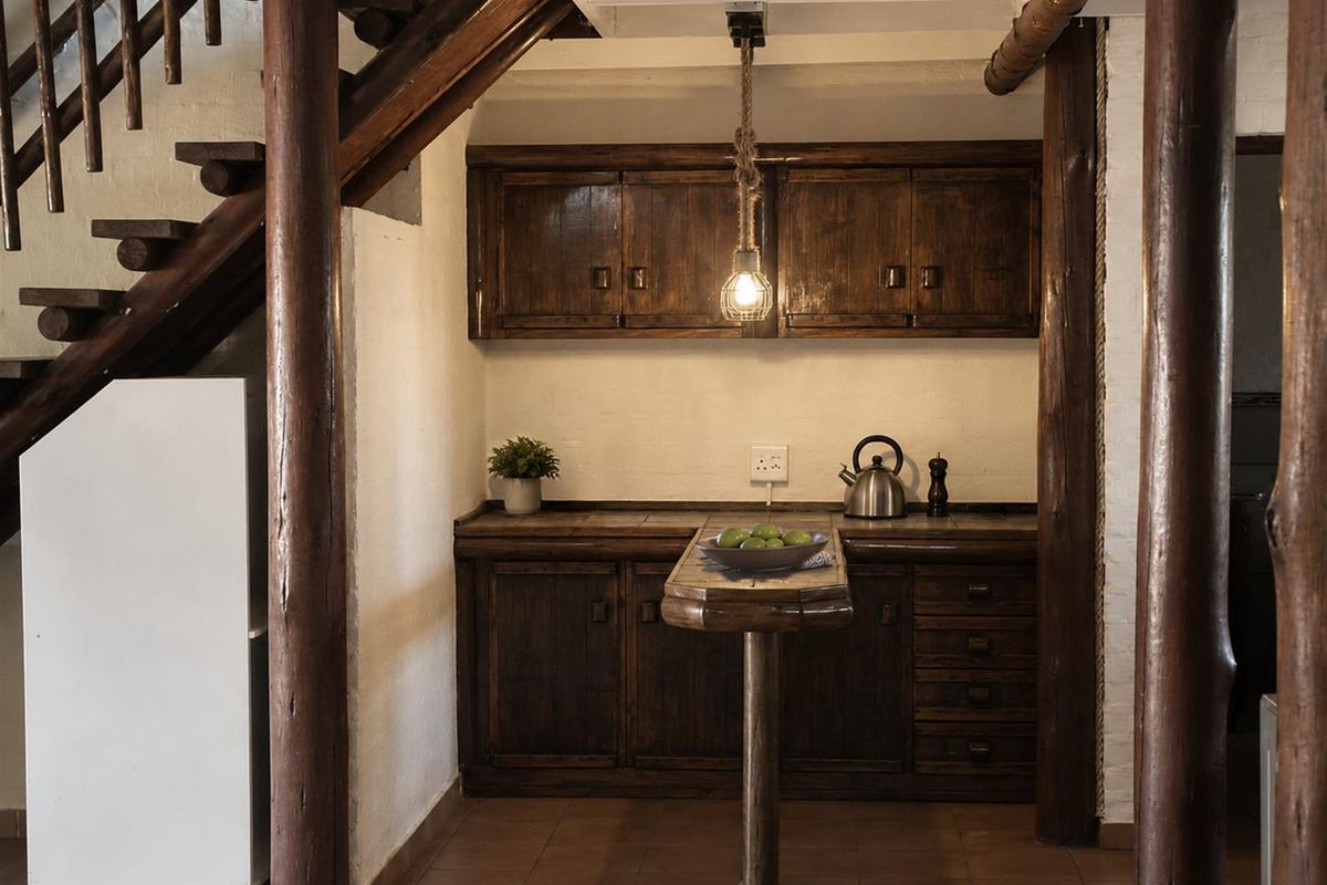 A rustic kitchen area featuring dark timber cabinetry, tiled countertops, and exposed wooden structural pillars, situated beneath a wooden staircase.