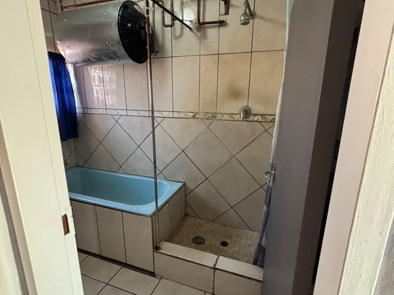 A dated bathroom featuring a blue bathtub, shower area, and exposed plumbing with an overhead water heater unit.