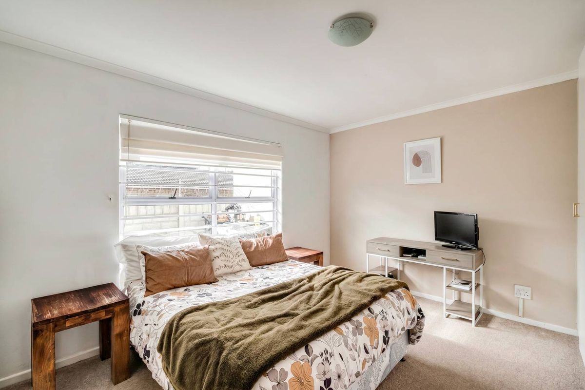 A well-lit bedroom featuring neutral wall tones, carpeted flooring, and a large window with fitted blinds.