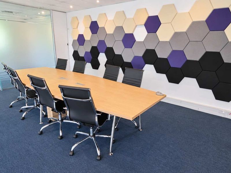 Modern office meeting room featuring glass partitions, acoustic hexagonal wall panels, recessed lighting, and professional carpet flooring.
