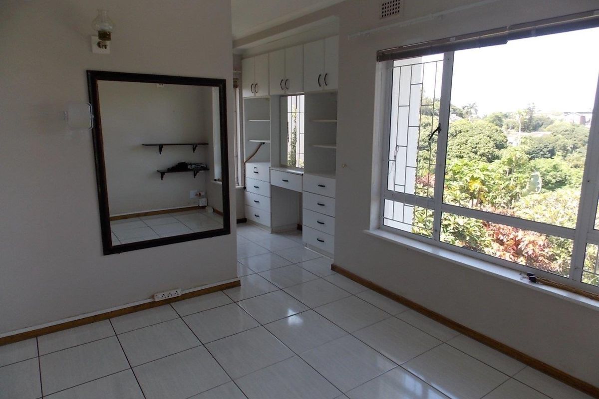 A bright bedroom featuring built-in cabinetry, a vanity area, tiled flooring, and large windows with security bars overlooking the surrounding greenery.