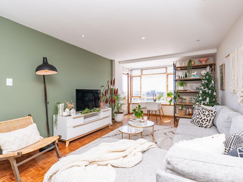 A bright and inviting living room featuring warm parquet flooring, a feature green wall, and large windows that provide ample natural light.