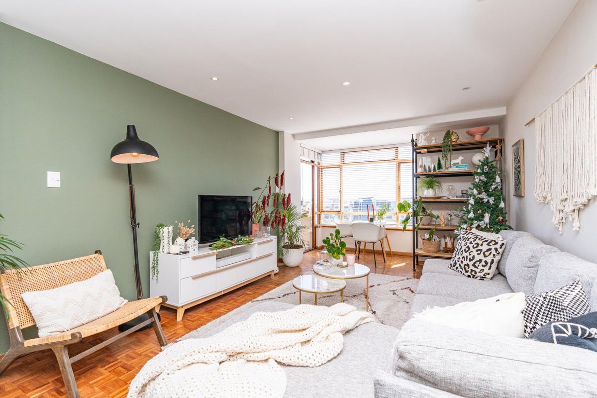 A bright and inviting living room featuring warm parquet flooring, a feature green wall, and large windows that provide ample natural light.