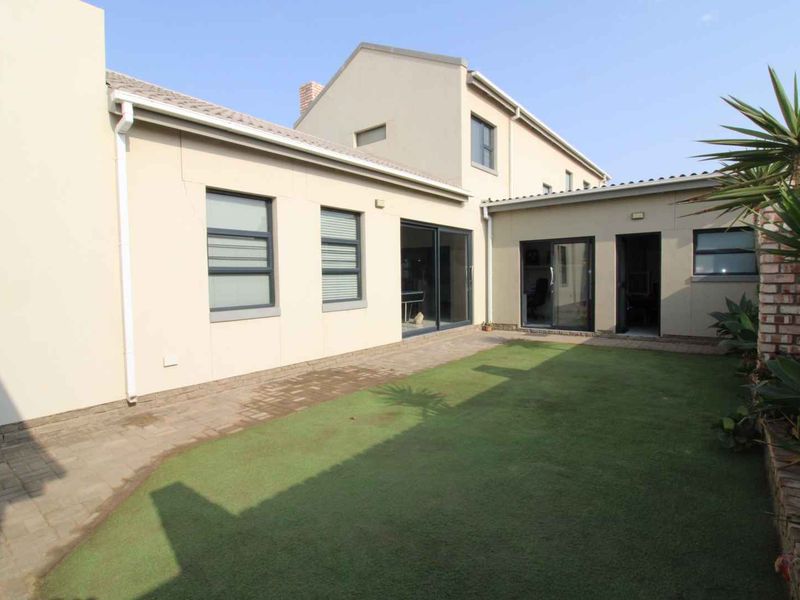 A well-maintained rear exterior view featuring a paved walkway, artificial lawn, and modern sliding glass doors providing access to the home.