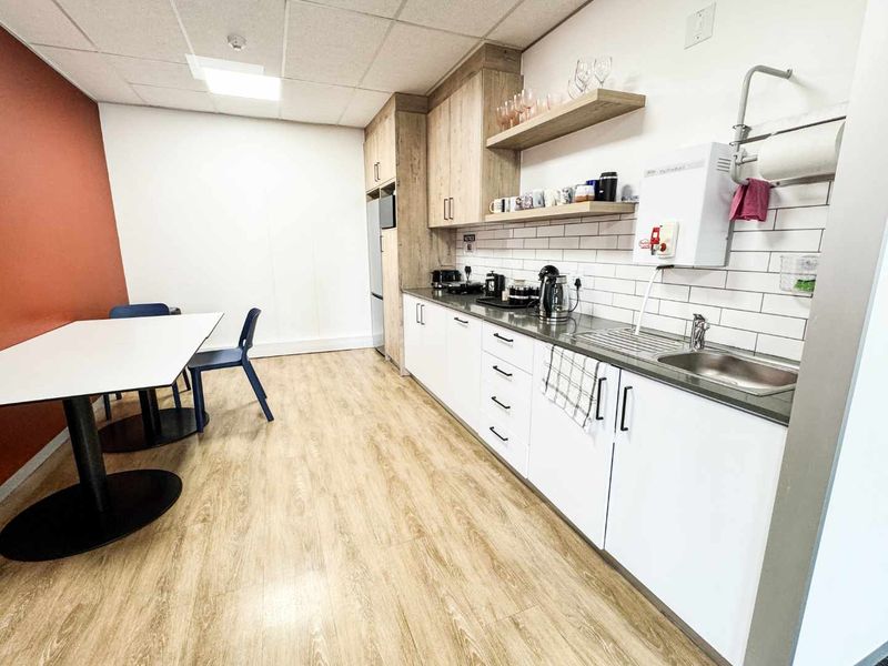 A clean and functional office kitchenette featuring modern white cabinetry, wood-look laminate flooring, subway tile backsplash, and a convenient instant hot water dispenser.