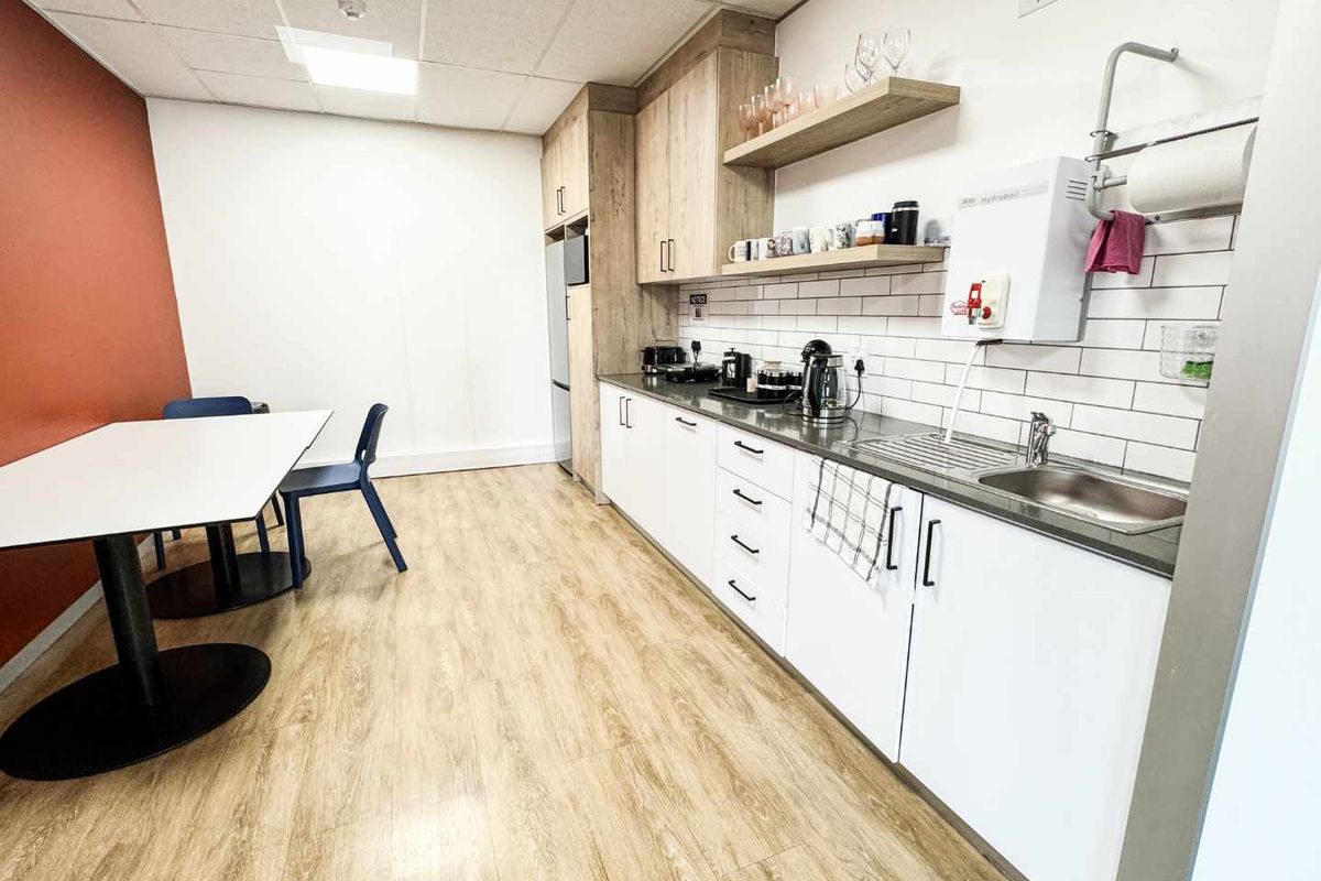 A clean and functional office kitchenette featuring modern white cabinetry, wood-look laminate flooring, subway tile backsplash, and a convenient instant hot water dispenser.