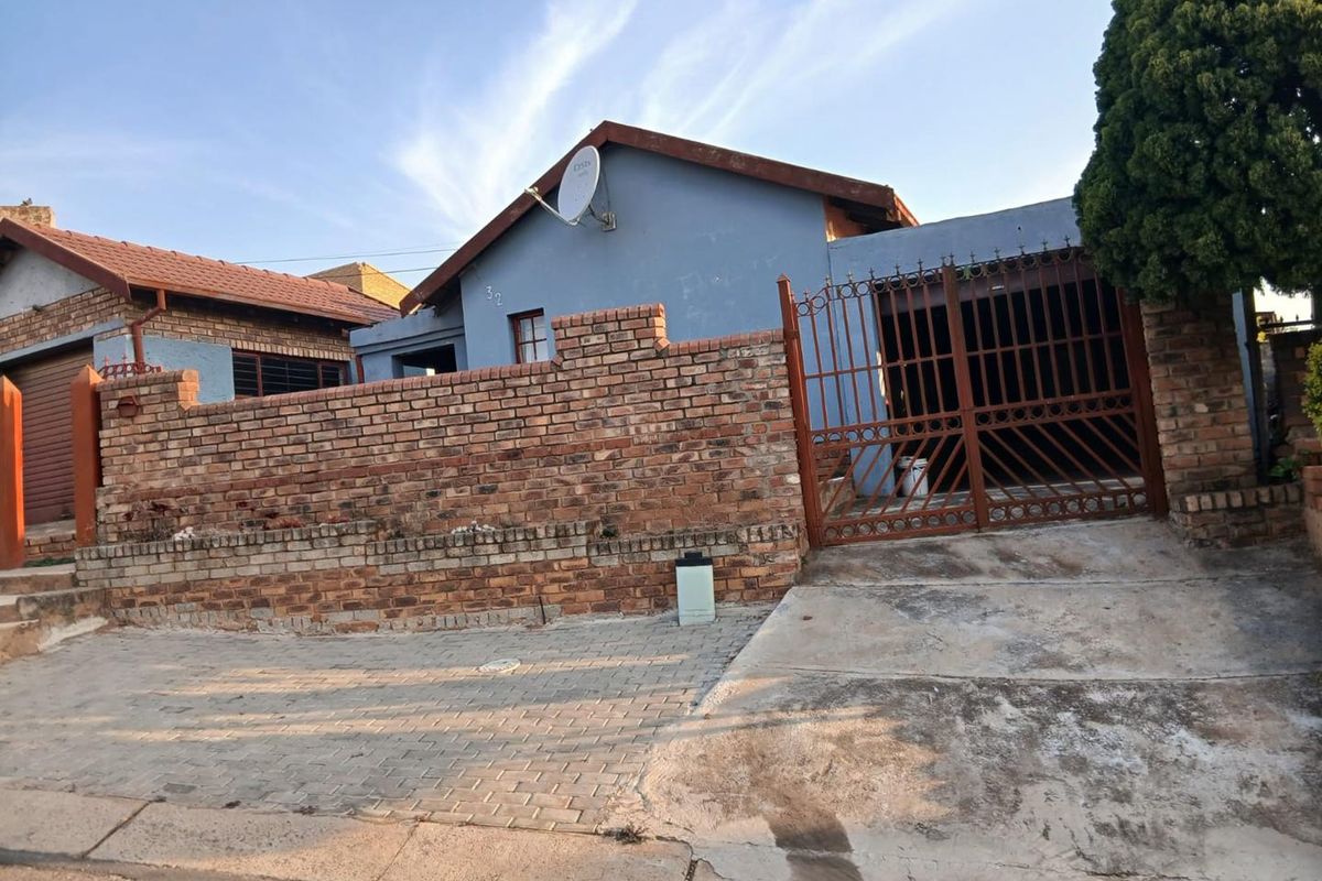 Front exterior view of a single-story house featuring a brick boundary wall, metal driveway gate, and paved driveway area.