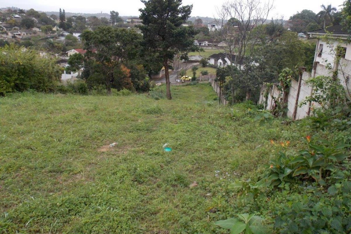 A sloped vacant residential plot featuring established trees and a precast concrete boundary wall, overlooking the surrounding neighborhood.