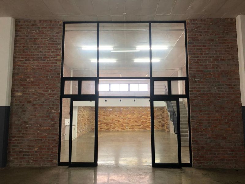 Spacious open-plan industrial style interior featuring exposed brick walls, polished concrete floors, and a prominent black steel-framed glass partition with an internal staircase.