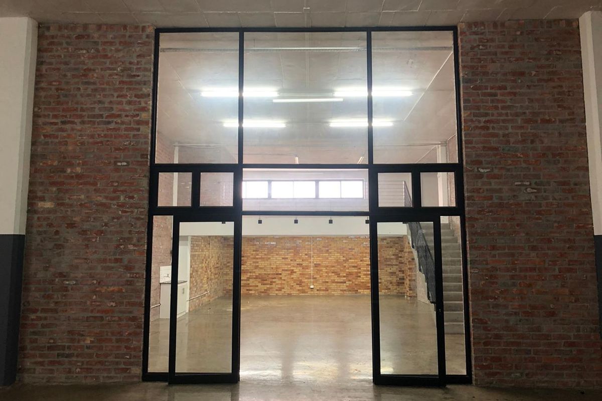Spacious open-plan industrial style interior featuring exposed brick walls, polished concrete floors, and a prominent black steel-framed glass partition with an internal staircase.