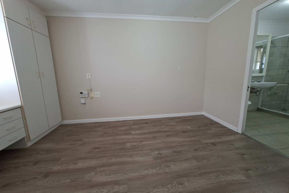 A bright, empty bedroom featuring modern wood-look laminate flooring, built-in cabinetry, and an adjoining en-suite bathroom.