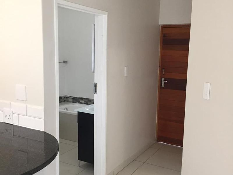 A clean, neutral-toned hallway featuring tiled flooring, a wooden entrance door, and a partial view into an adjacent bathroom and kitchen area.