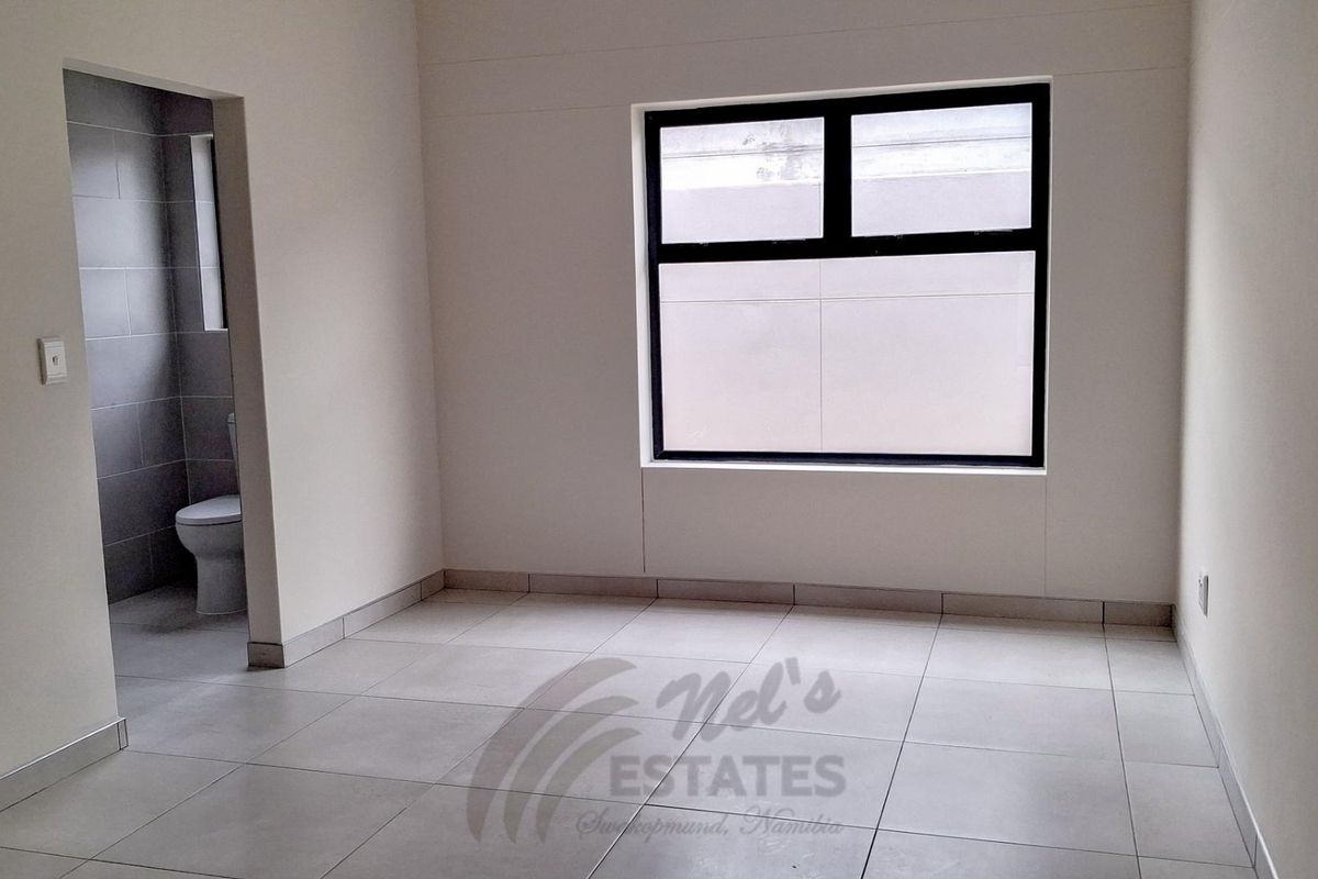 A bright, empty bedroom featuring neutral tiled flooring, modern black-framed windows, and an adjoining en-suite bathroom.
