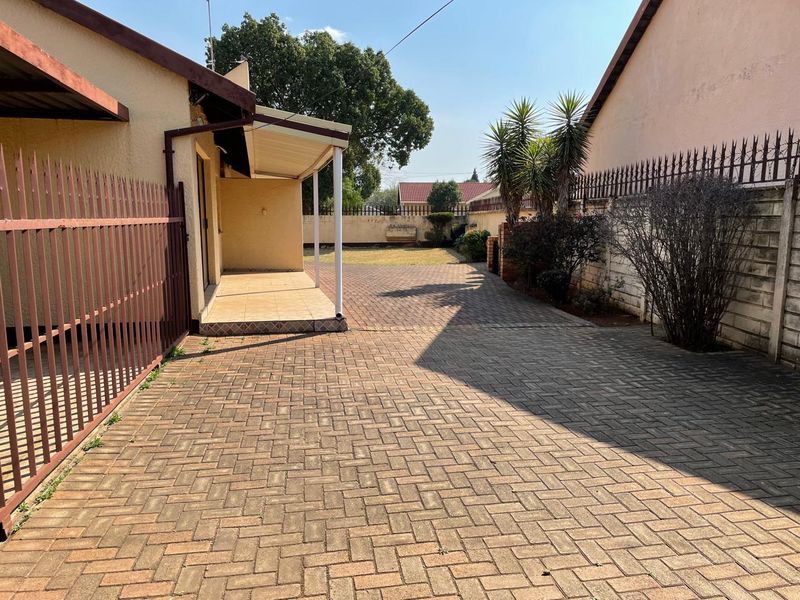 A rear exterior view featuring a paved driveway, a covered patio area, and secure perimeter walling with metal fencing.