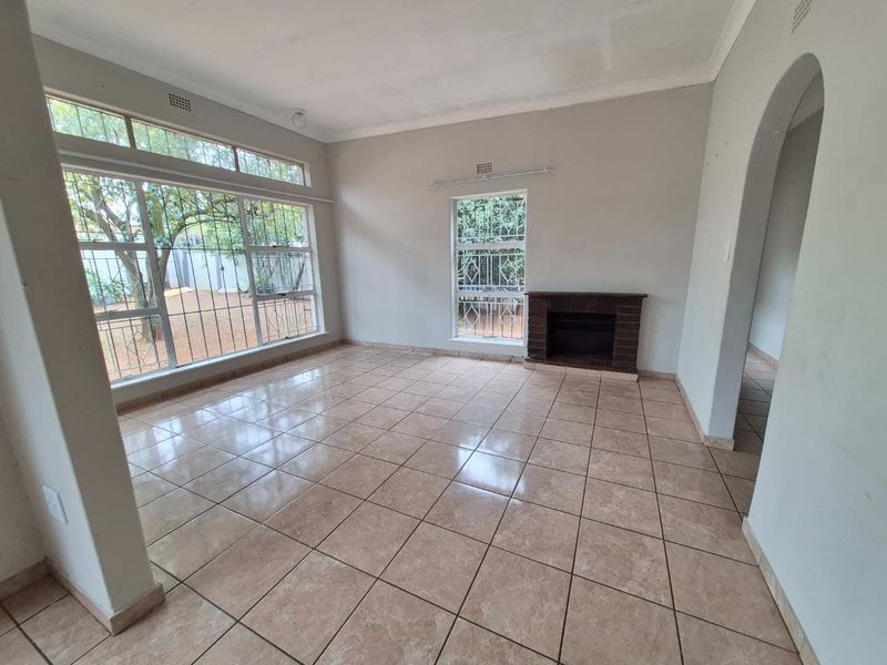 A spacious, empty living room featuring tiled flooring, a brick fireplace, and large windows with security bars, offering a blank canvas for interior design.